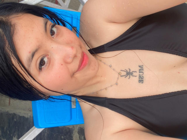 Elizaa, con el cabello negro y húmedo, ojos marrones, un piercing en la nariz y una camiseta negra que deja ver un tatuaje de araña en su pecho. Está sentada en una silla azul, mirando a la cámara..