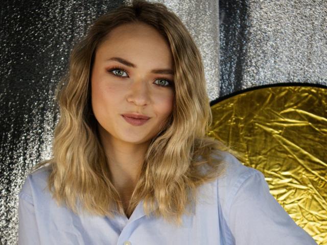 SallyVerda with blonde wavy hair, wearing a white shirt, has green eyes, light makeup, and a subtle smile, standing in front of a textured silver and gold background.