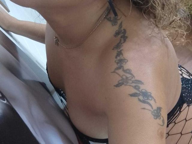 SolmaryHot's tattooed shoulder, featuring a floral design, with curly blonde hair, wearing black fishnet lingerie and a gold necklace, showing her cleavage and upper chest.