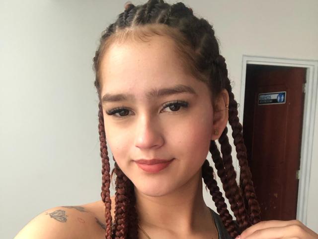 Melody Mansson a les cheveux bruns tressés, une peau claire, un petit tatouage sur l'épaule, un maquillage discret et un sourire charmant. Ses petites seins sont à peine visibles dans le cadre..