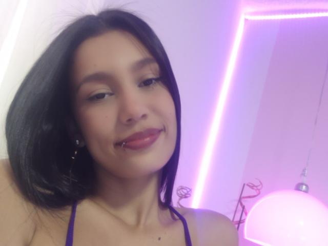 Kataleya Mansson, com cabelo preto, olhos castanhos e um piercing no lábio, sorrindo com um pequeno piercing no nariz, vestindo uma blusa roxa, em um quarto com iluminação suave e luzes neon rosa..