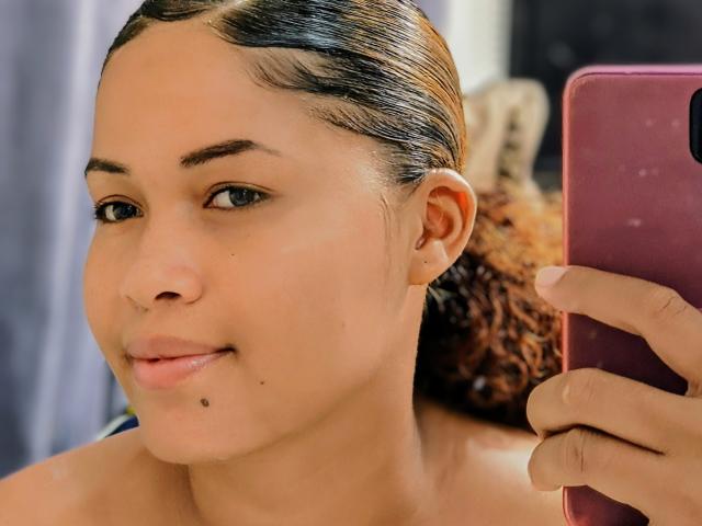 IrisI69 with light brown skin, black eyebrows, small mole on chin, curly brown hair, holding pink phone, taking selfie, smiling, slightly parted lips, background blurred.
