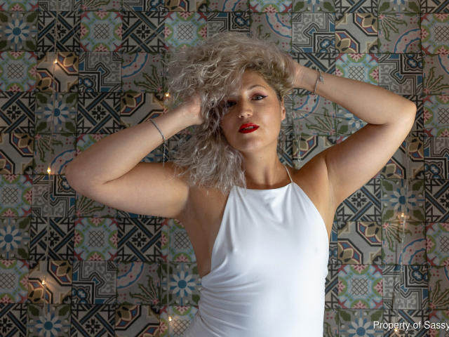 SassyKate with curly grey hair, red lipstick, white halter dress, hands in hair, exposing armpits, small tattoos on wrists, standing, confident, sexy.