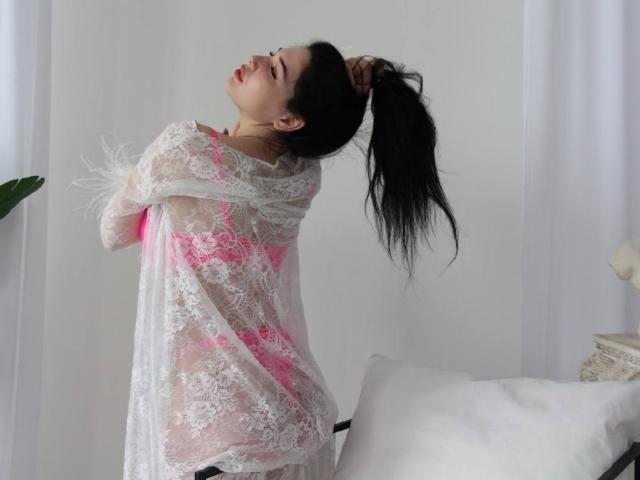 LiyaFrench with black hair in a ponytail, wearing a sheer white lace dress with pink accents, holding her hair up, eyes closed, facing left, in a white room.