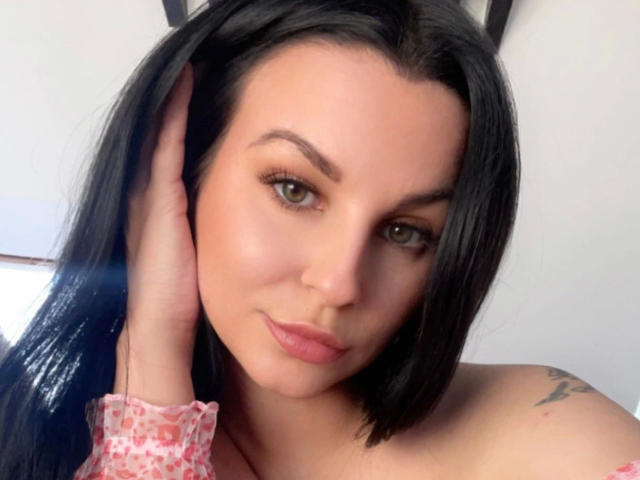 Kyra-Banx-hot has black hair, green eyes, and is touching her head with a pink, patterned sleeve. Her tattoo is on her right shoulder, and her tits are exposed.