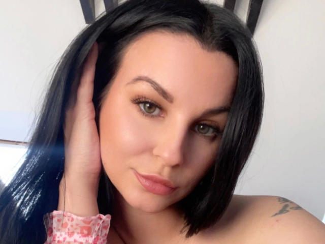 Kyra-Banx-hot has black hair, green eyes, and a slight smile. Her hand is on her head, showing a pink, patterned wristband. She has a tattoo on her right shoulder.
