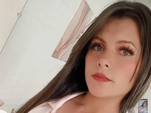 VictoriaWaynes has long brown hair, green eyes, and pink lips, wearing a pink shirt, with visible eyelashes and a subtle smile. Her tits are not visible.