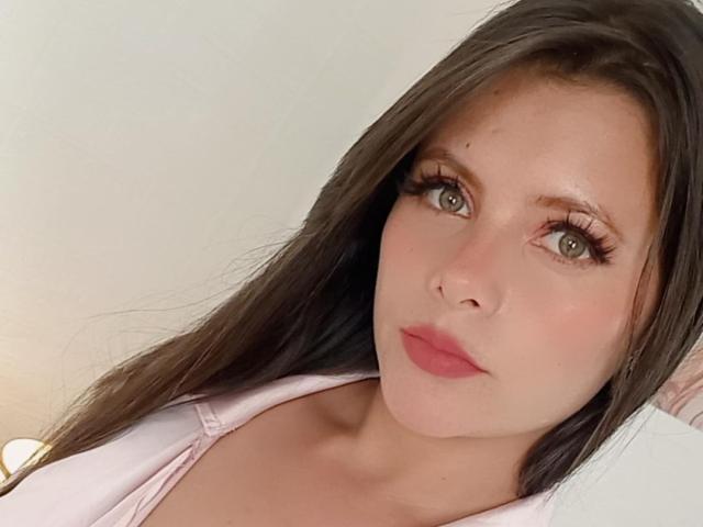 VictoriaWaynes has long brown hair, green eyes, and pink lips. She's wearing a pink top, showing her small boobs and smooth skin. Her makeup includes long, dark eyelashes.