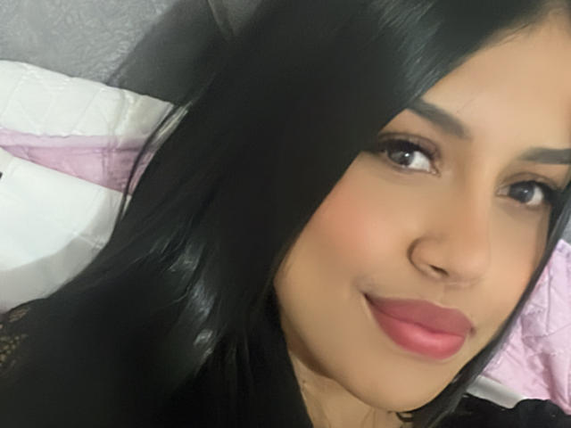 StephanieDavies has long black hair, brown eyes, and pink lipstick. She's smiling with a slight nose and visible black eyebrows. She's wearing a black top.