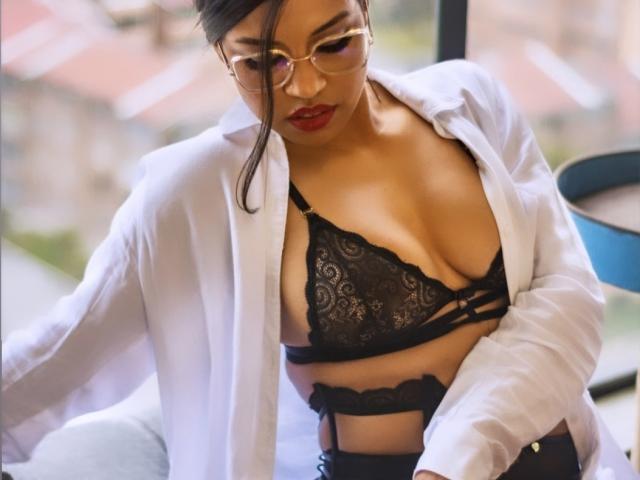 Stephanie Davies, con lápiz labial rojo, gafas, camisa blanca abierta, un sujetador de encaje negro que deja ver sus grandes senos, y una liga negra, mirando hacia abajo, en una habitación iluminada por el sol, con una silla azul..
