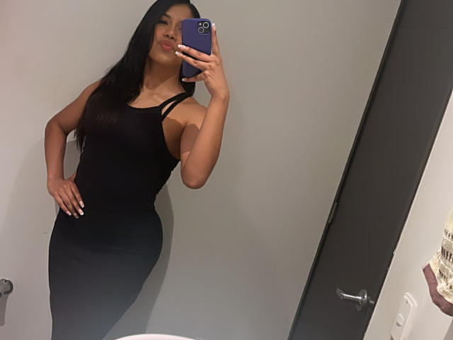 Stephanie Davies, con un vestido negro ajustado que muestra su trasero y sus pequeños senos, se toma un selfie frente al espejo con un teléfono morado, tiene el pelo largo y negro, una mano en la cadera y está sonriendo..