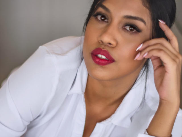 Stephanie Davies tiene la piel morena, ojos oscuros, lápiz labial rojo y lleva una camisa blanca, con una mano tocándose el pelo, mostrando uñas rosas. Sus senos no son visibles..