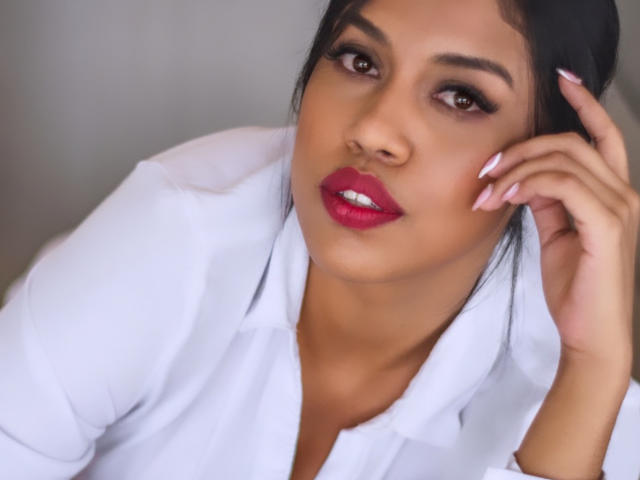 Stephanie Davies, con cabello oscuro, ojos marrones, lápiz labial rojo, camisa blanca y uñas rosas, tocándose la cabeza con una expresión seria, mostrando su rostro sexy y atractivo..