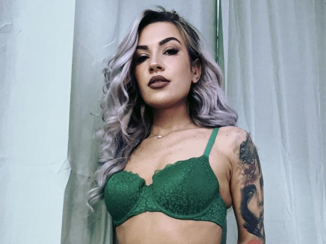 ZoeyMarie with gray wavy hair, green lace bra, showing small tits, tattooed arm, and a neutral expression, standing in front of a curtain.