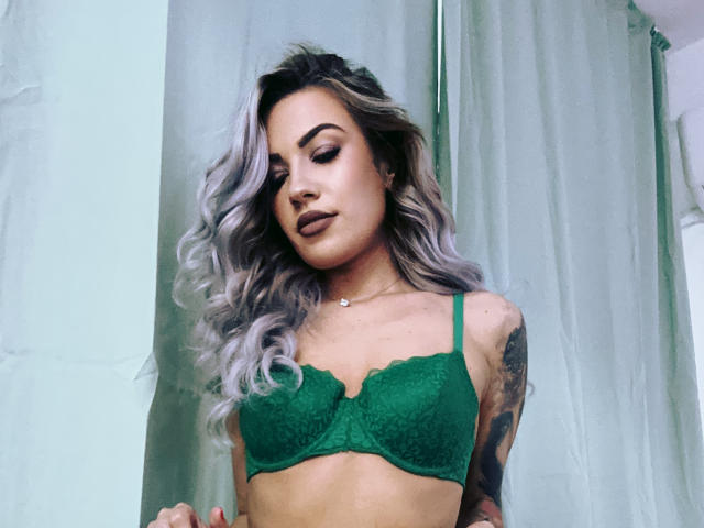 ZoeyMarie with wavy gray hair, green lace bra, tattoos on right arm, and dark lipstick, looking down, standing in front of green curtains. Boobs, cute, sexy.