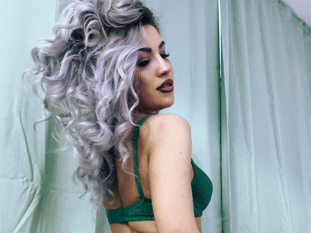 ZoeyMarie mit lockigem, silberfarbenem Haar, einem grünen BH und dunklem Lippenstift, blickt über ihre Schulter und zeigt ihre Brüste. Sie steht vor einem hellgrünen Vorhang..