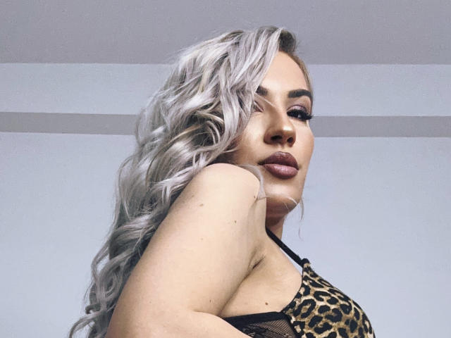 ZoeyMarie with long, wavy, silver hair, wearing a leopard print top, showing her boobs, with dark makeup and a confident, sultry expression.