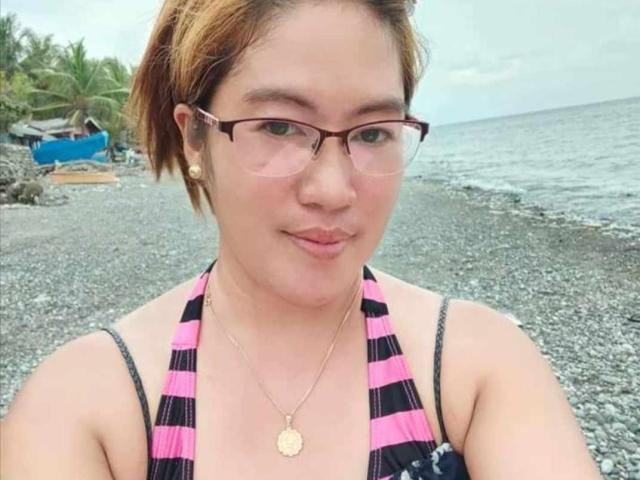 Xenia69 with short brown hair, glasses, and pink-striped bikini top, showing her small tits, wearing a gold necklace, standing on a pebbly beach, smiling slightly.