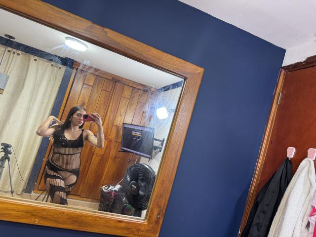 SharonKlart in zwarte visnetlingerie met grote borsten, die een rode telefoon vasthoudt en een spiegelzelfportret maakt in een kamer met blauwe muren. Houten spiegelomlijsting, ventilator en gordijn op de achtergrond..