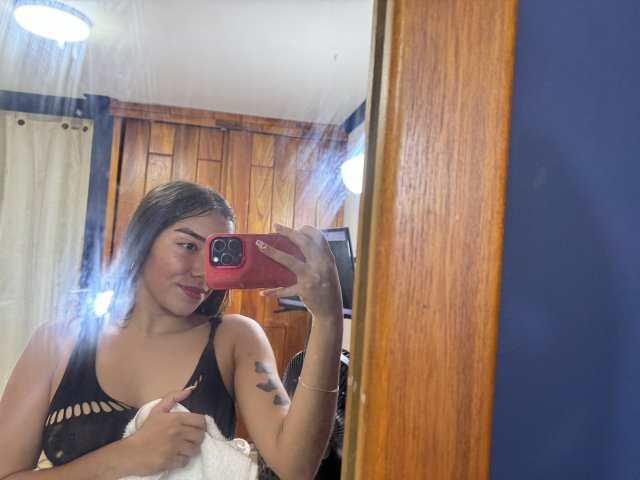 SharonKlart, avec ses cheveux foncés, portant un haut en maille noire, tenant une serviette, prenant un selfie devant un miroir avec un téléphone rouge, montrant un tatouage sur son bras, souriante..
