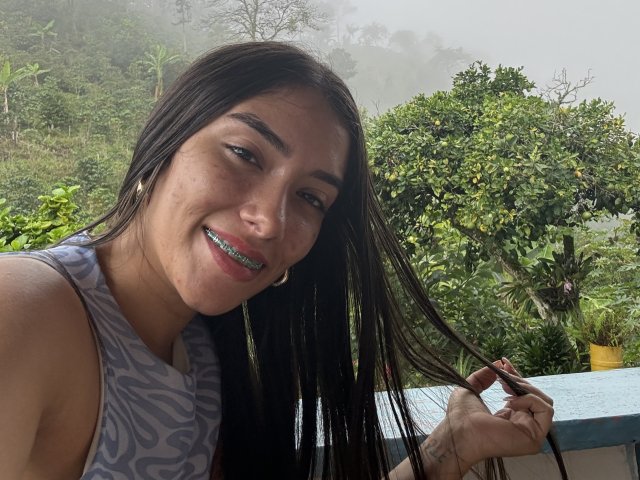 SharonKlart with long dark hair, smiling, holding a lock, wearing a patterned sleeveless top, visible teeth with green braces, standing near a blue railing, foggy background.