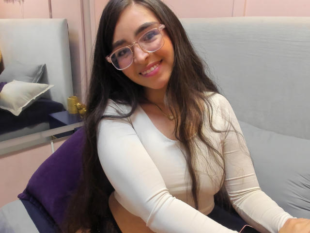 EvaMcAllister with long black hair, glasses, and a white long-sleeve top, smiling at the camera, showing her small tits and a cute face. She's sitting on a bed.