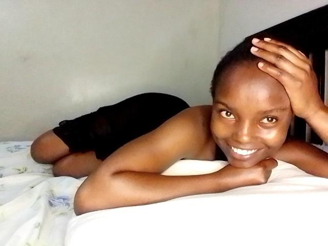 Africanah, di carnagione scura, sorridente, sdraiata sul letto, vestito nero, spalle scoperte, mano sulla testa, lenzuolo bianco, sorride alla fotocamera, occhi marroni, capelli neri corti, rilassata, carina..