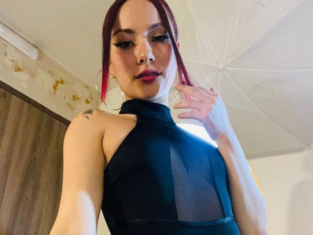 ValeryHarllow with red hair, wearing a black sheer top, showing her tits, hand near face, slight smile, tattoo on left shoulder, ceiling fan above.