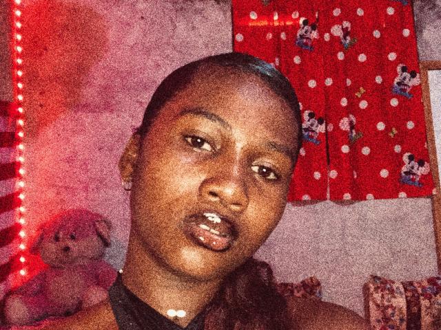 Jobby's dark brown face, glossy lips, and short hair, wearing a black choker, in a grainy, red-lit selfie with a stuffed pink rabbit in the background.
