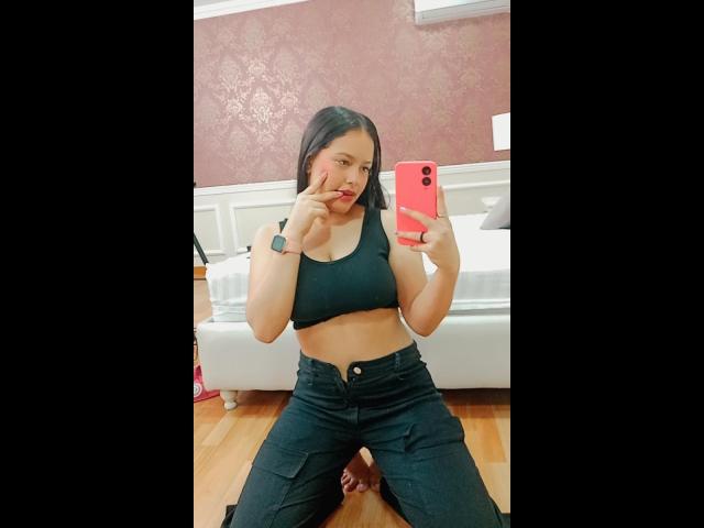 LaurenMartins with black crop top, showing tits, and black pants, kneeling, making a peace sign, holding a pink phone, wearing a black wristwatch, in a bedroom.