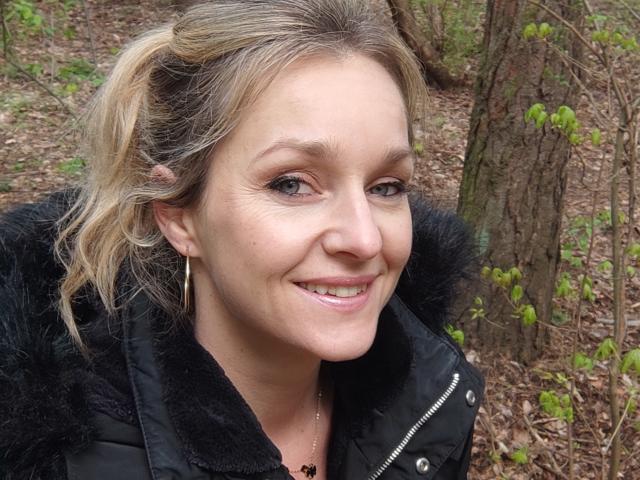 AliciaSecret, blonde, souriante, avec des yeux verts, veste noire, petites boucles d'oreilles, queue de cheval légèrement ébouriffée, peau claire, lèvres roses, fond en bois avec des arbres et des feuilles vertes..
