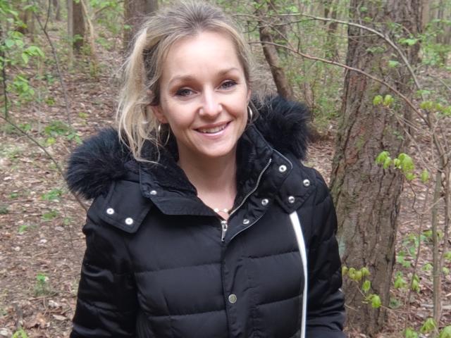AliciaSecret hat blondes Haar, blaue Augen und ein Lächeln und trägt eine schwarze, gepolsterte Jacke mit einer schwarzen Fellhaube. Sie steht in einem bewaldeten Gebiet..