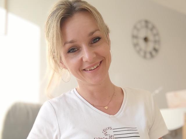 AliciaSecret, blonde, smiling, white tee, small tits, hoop earrings, necklace, light skin, casual, soft light, clock on wall, relaxed, attractive, upper body shot, slight tilt, happy.