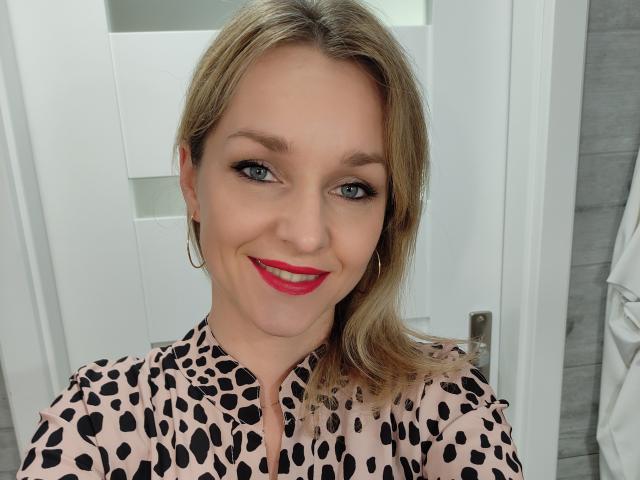 AliciaSecret mit blonden Haaren, blauen Augen und rotem Lippenstift, lächelnd in die Kamera, trägt ein Leopardenmuster-Shirt, Ohrringe im Hula-Hoop-Stil und zeigt ihr süßes Gesicht und ihren Hintern..