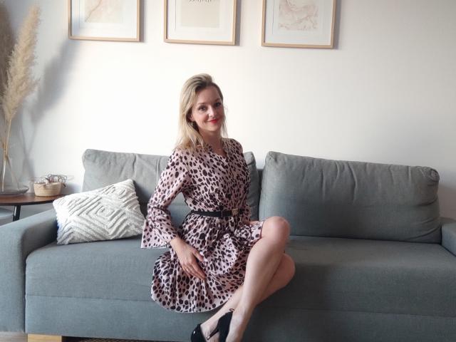 AliciaSecret, blonde, assise sur un canapé gris, portant une robe à motifs léopard, des talons noirs, les jambes croisées, un coussin blanc, une plante en pot, trois œuvres d'art encadrées sur un mur blanc..