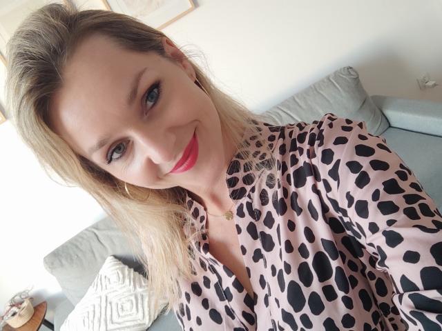 AliciaSecret with blonde hair, red lipstick, and a pink leopard print blouse, smiling at the camera, showing her tits slightly through the open shirt.