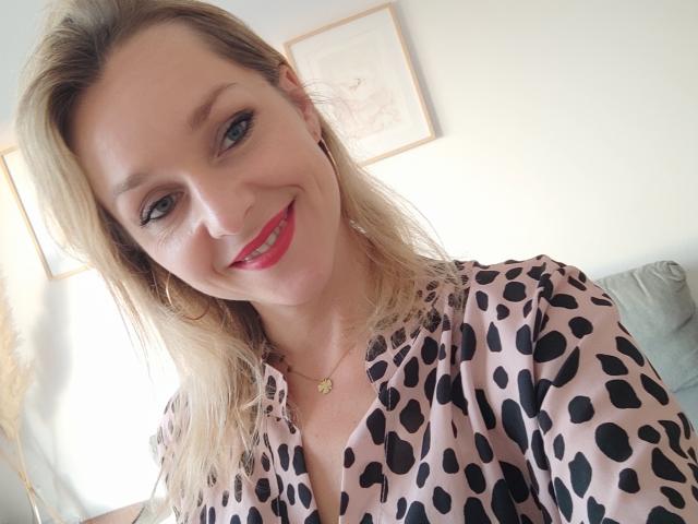 AliciaSecret with blonde hair, pink leopard print shirt, red lipstick, smiling, wearing gold necklace, hoop earrings, and a small star pendant, in a bright indoor setting.