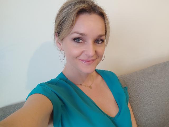 AliciaSecret with blonde hair, blue eyes, and a smile, wearing a teal top, hoop earrings, and a gold necklace, sitting on a gray couch. Her tits are visible.