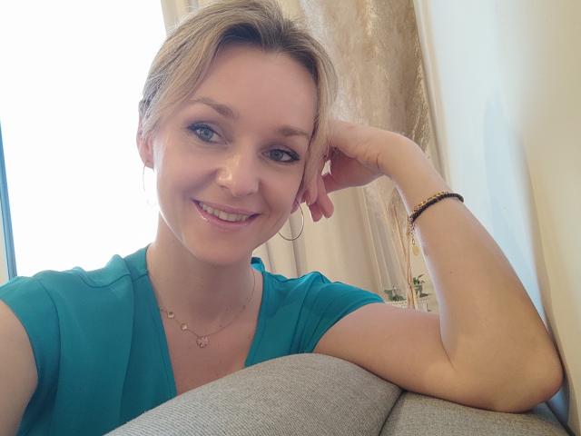 AliciaSecret, blonde, souriante, yeux bleus, chemise verte, main sur la tête, bracelet, collier, lumière douce, décontractée, mignonne, mur blanc, canapé gris, détendue, naturelle, confiante, légèrement rougie..