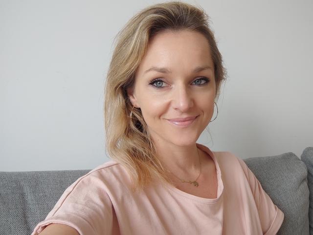 AliciaSecret with blonde hair, blue eyes, and a subtle smile, wearing a pink shirt, sitting on a gray couch, small gold necklace, slight makeup, and a relaxed expression.