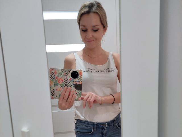 AliciaSecret with blonde hair, white tank top, small tits, blue jeans, holding a phone with a colorful pattern, smiling, reflected in a mirror, wearing hoop earrings.