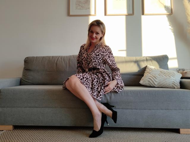 AliciaSecret, avec des cheveux blonds, portant une robe à motifs léopard, des talons noirs, assise sur un canapé gris, souriante, montrant ses fesses, les jambes croisées, la lumière venant de la droite..
