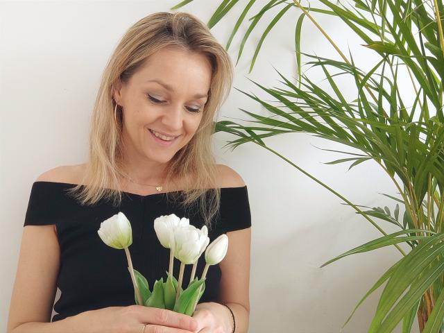 AliciaSecret, blonde, smiling, holding white tulips, black off-shoulder top, green plant to the right, white wall background, light skin, subtle makeup, casual, cute.