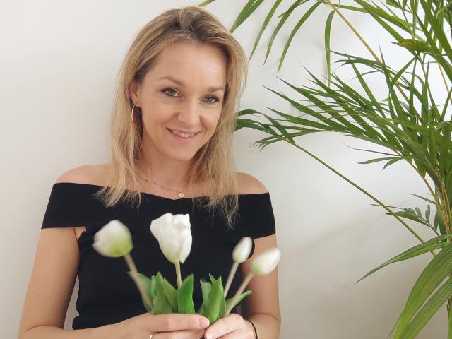 AliciaSecret with blonde hair, off-shoulder black top, holding white tulips, smiling, green plant to the right, fair skin, subtle makeup, and a small gold necklace.