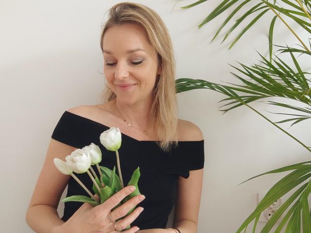 AliciaSecret, cheveux blonds, robe noire à épaule dénudée, tenant des tulipes blanches, sourire discret, feuilles de plantes vertes, maquillage léger, décontractée, élégante, éclairage doux, expression douce, mignonne, détendue..