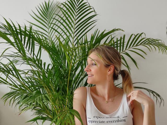 AliciaSecret, avec une queue de cheval blonde, un débardeur blanc et un collier, se tient à côté de feuilles de palmier vertes, regardant sur le côté avec un léger sourire. Ses seins sont petits et mignons..