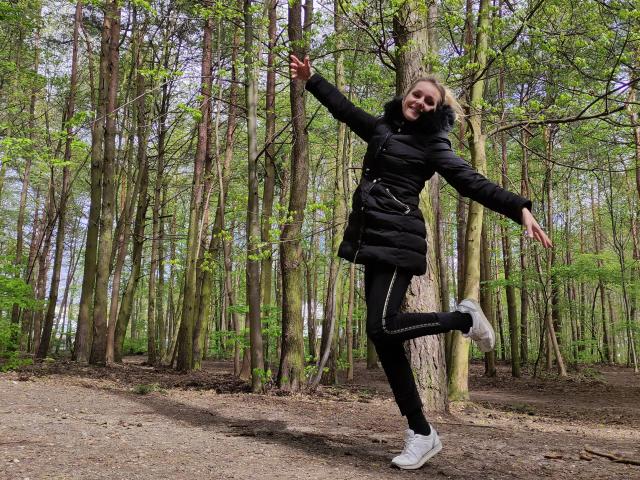 AliciaSecret, black puffy jacket, black pants, white sneakers, jumping in forest, arms out, legs bent, smiling, curly brown hair, tree trunks behind, dirt path below.