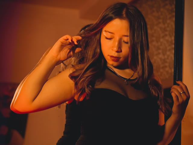 NatashaBlank with wavy brown hair, wearing a black dress with a sheer neckline, touching her hair, standing in warm orange light, eyes closed, wearing a necklace.