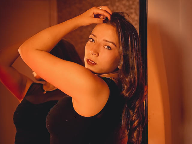 NatashaBlank with dark brown hair, wearing a black sleeveless top, raising her arm, showing her tits in a mirror, has a ring on her finger, and a sexy look.
