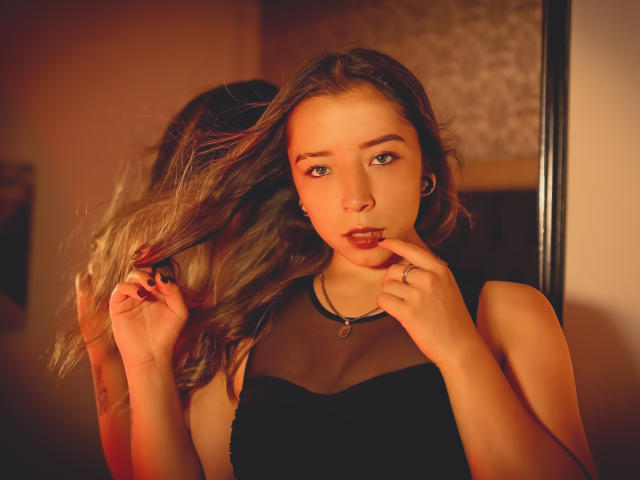 NatashaBlank with wavy brown hair, blue eyes, and red lipstick, wearing a black dress, touching her lips with fingers, reflection in mirror, warm orange lighting.